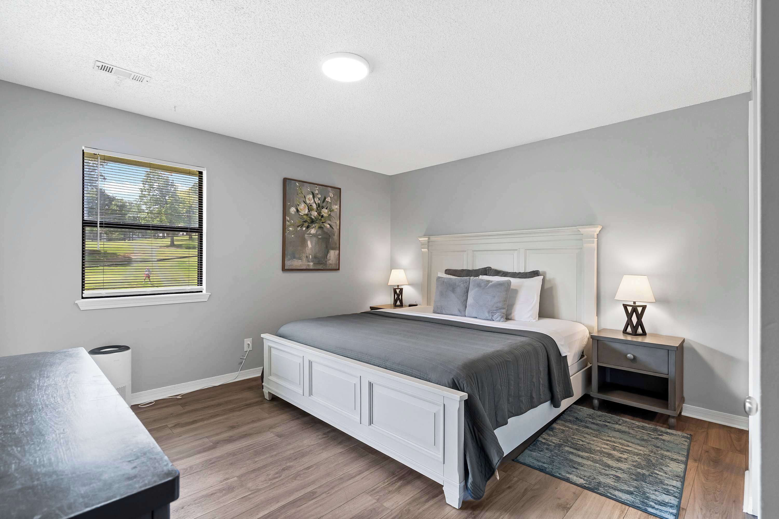 A bedroom with a bed, bedside tables, a window with blinds, a painting, and a lamp.