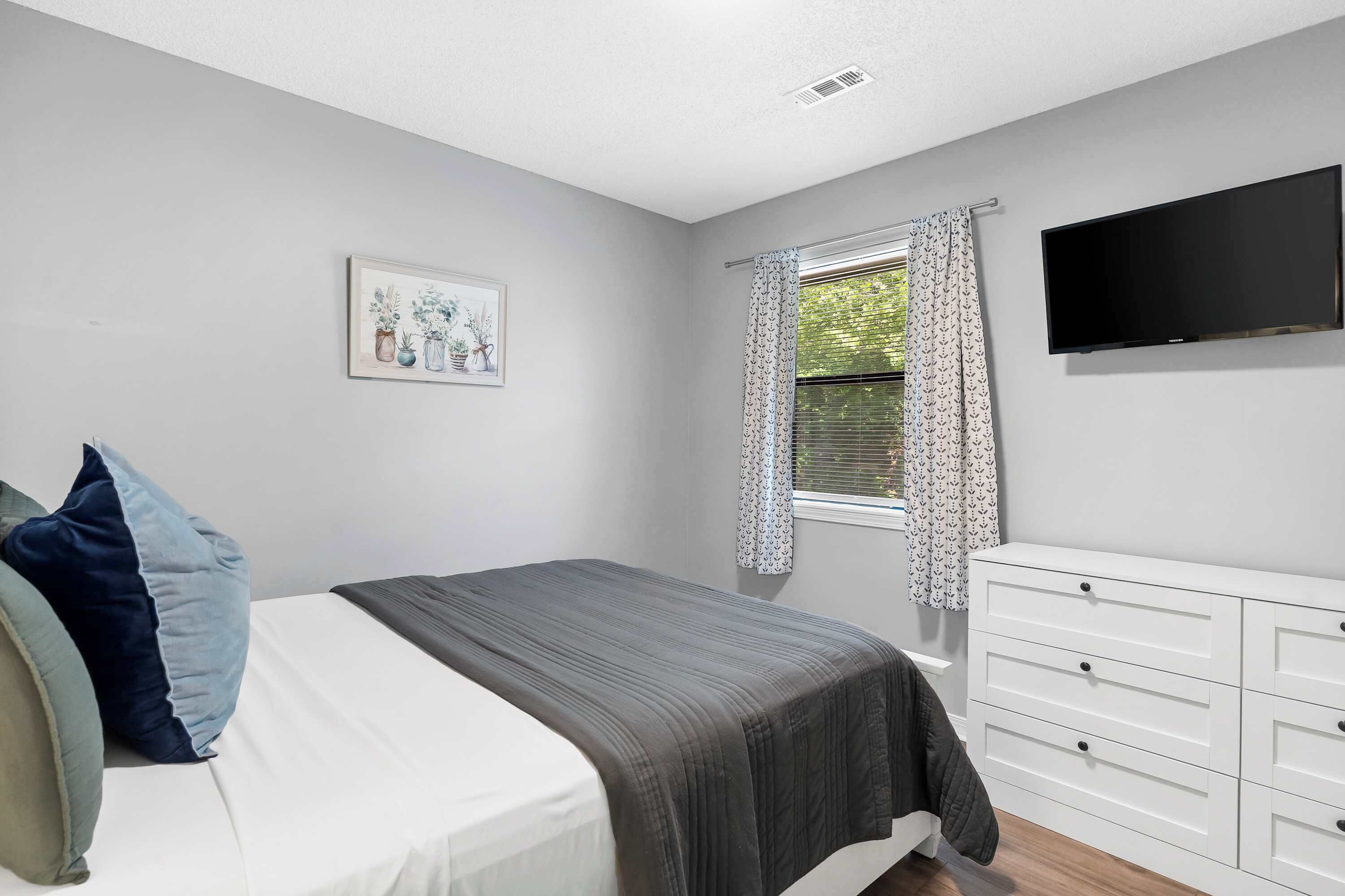 A bedroom with a bed, a dresser, a television, and a window with curtains.