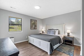 A bedroom with a bed, bedside tables, a window with blinds, a painting, and a lamp.
