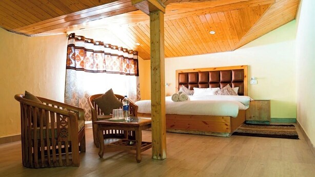 A bedroom with a wooden headboard, a wooden table with a chair, and a window with patterned curtains. - Shimla Hill View (Shimla)