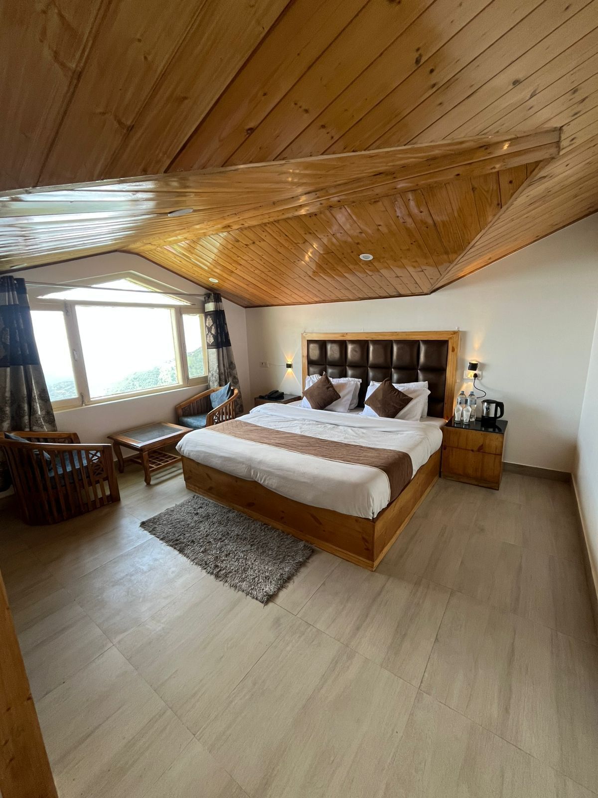Quarto com teto de madeira, uma cama grande com cabeceira, mesas de cabeceira, janela com cortinas e vista para o exterior.