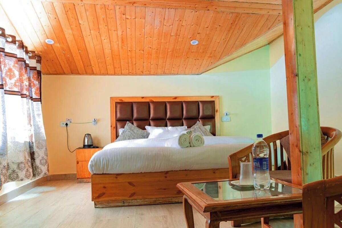 A bedroom with a wooden headboard, a bed with pillows, a nightstand with a kettle, and a glass table with a bottle.