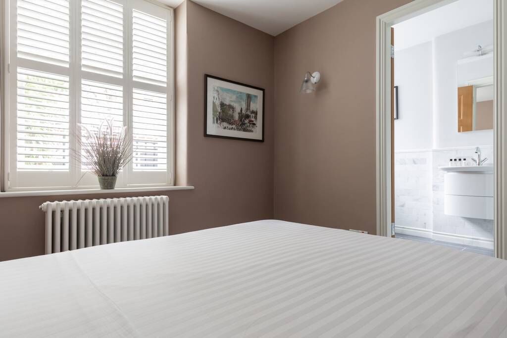 Un dormitorio con una cama grande, un radiador, una ventana con persianas, un cuadro enmarcado en la pared y un baño visible a través de una puerta abierta.