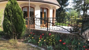 A building with a circular balcony, surrounded by a garden with various flowers and a tall evergreen tree.