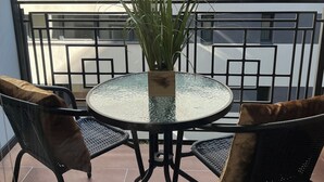 A round glass table with a potted plant, surrounded by wicker chairs with brown cushions, on a balcony with a metal railing and a building in the background.