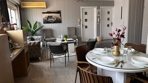 En matplats med ett runt bord i glas, dukat för fyra personer, omgivet av stolar i rotting. En kristallkrona hänger från taket, och en krukväxt står på bordet.