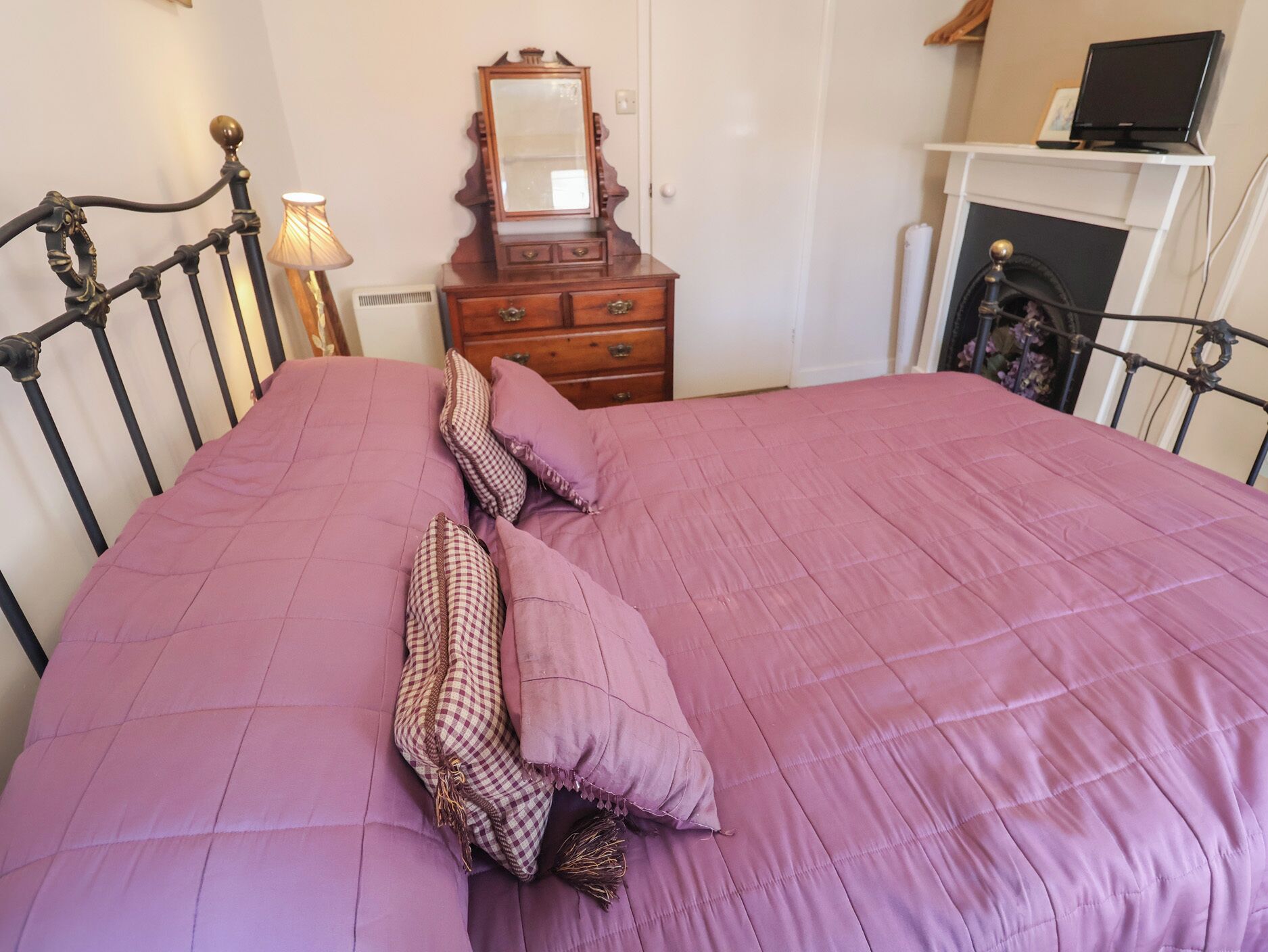 A bed with a purple quilt and pillows, a wooden dresser with a mirror, and a fireplace with a TV on top.