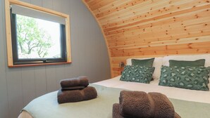 Una camera accogliente con un soffitto in legno curvo, un'ampia finestra con vista sugli alberi, un letto rifatto con cuscini verdi e marroni e due asciugamani arrotolati sul letto.