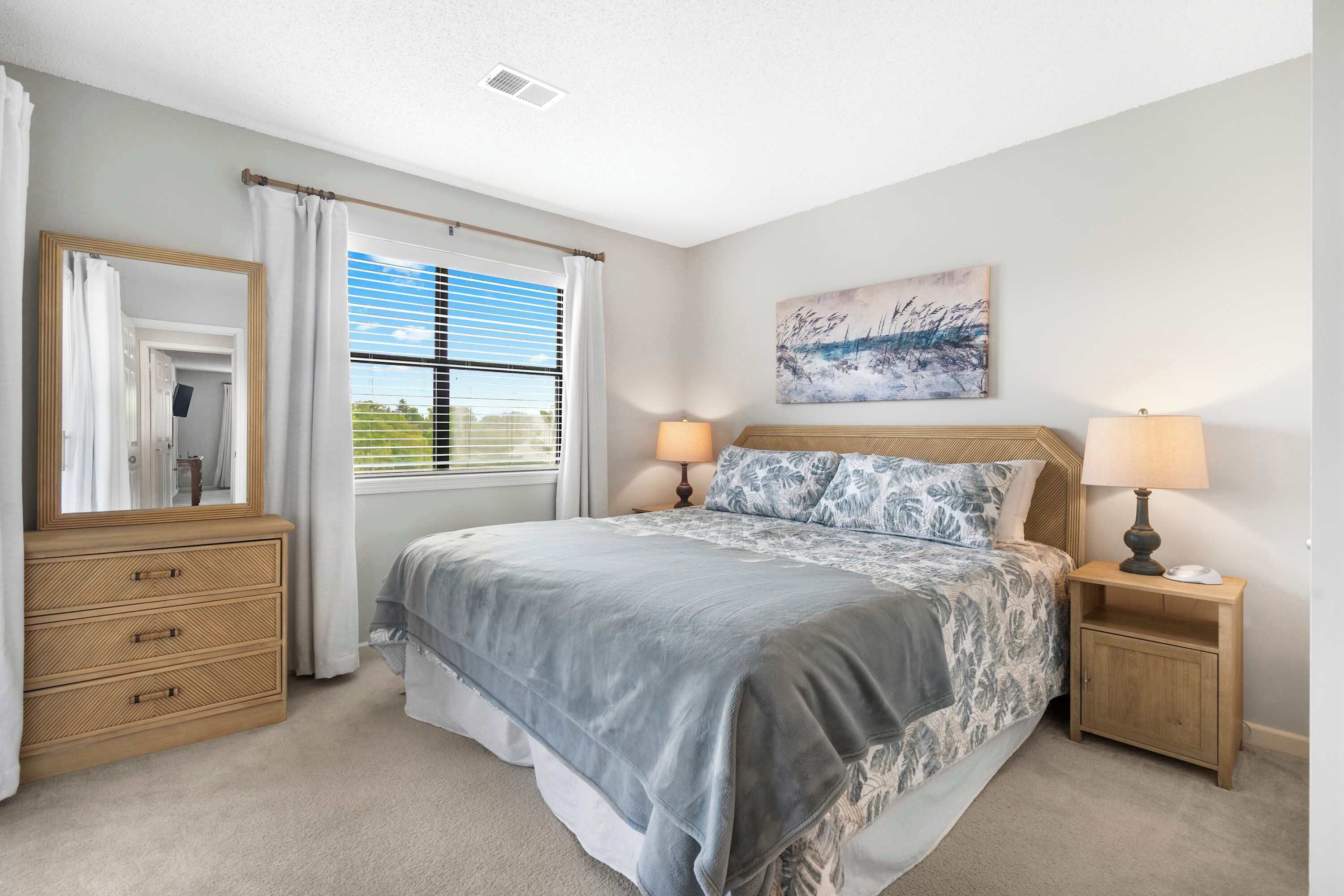 A bedroom with a large bed, two bedside tables with lamps, a dresser, a window with blinds, and a painting on the wall.