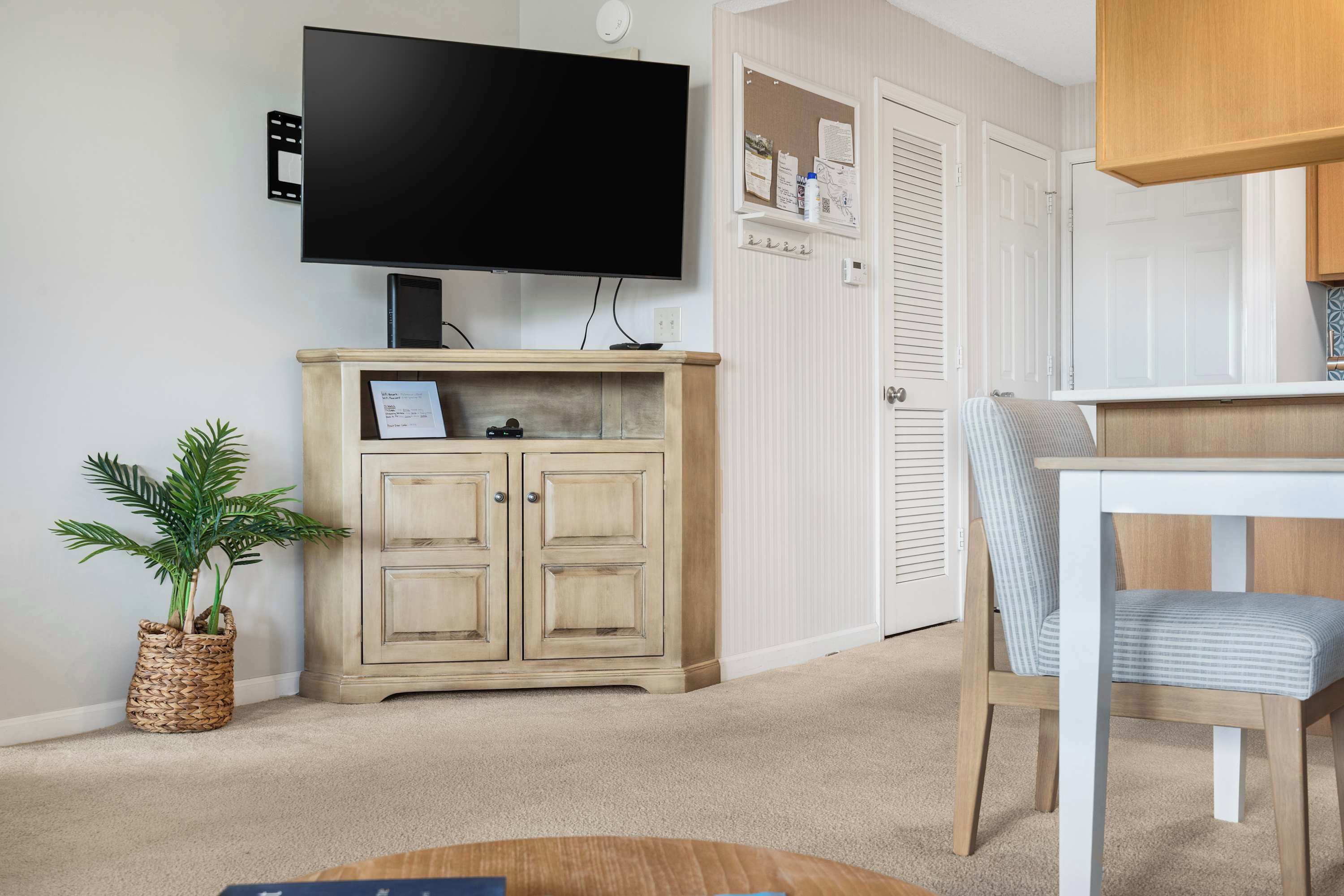 A living room with a flat-screen TV mounted on the wall, a wooden cabinet with a picture frame, a potted plant, and a dining table with a chair.