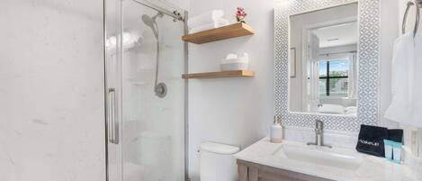 A modern bathroom with a glass shower enclosure, a white toilet, wooden shelves above the sink, and a large mirror with built-in lights.