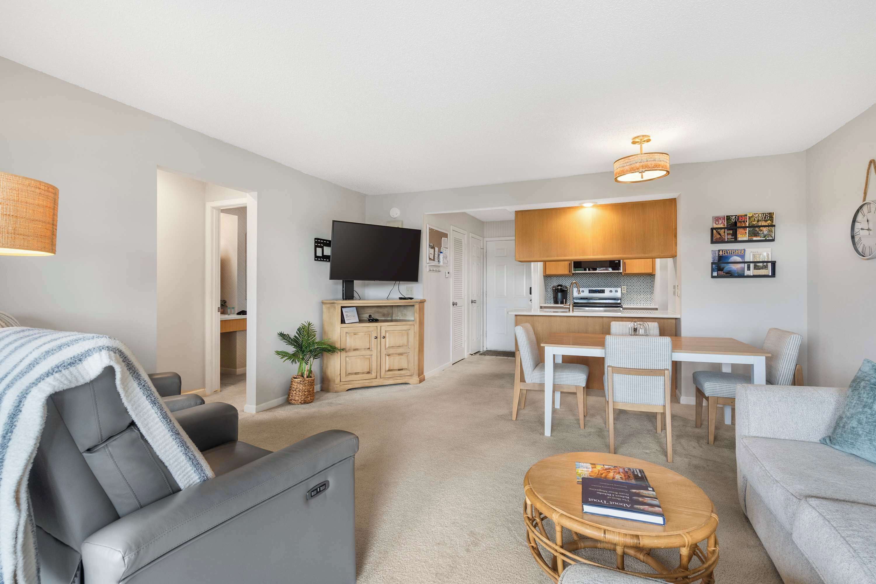 A modern living room with a grey sofa, a coffee table with a book, a dining area with a wooden table and chairs, and a kitchen with a TV and cabinets.