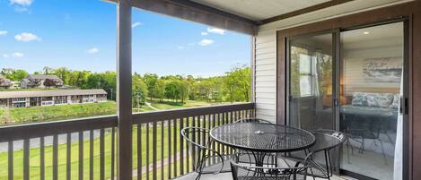 A balcony with a metal table and chairs, a sliding glass door, and a view of a green landscape with buildings in the distance.