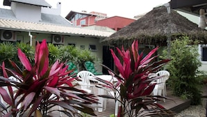 A patio area with a thatched roof, surrounded by potted plants and white plastic chairs. - Pousada Aloha Mariscal (Bombinhas)