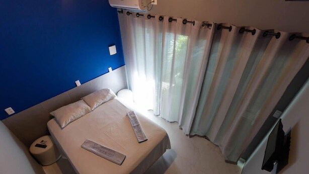 A bedroom with a bed, a bedside table, a wall-mounted air conditioner, and a window with curtains. - Pousada Aloha Mariscal (Bombinhas)