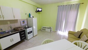 A compact kitchen with white cabinets, a stove, and a refrigerator. There is a dining table with chairs and a sofa in the living area. A television is mounted on the wall. - Pousada Aloha Mariscal (Bombinhas)