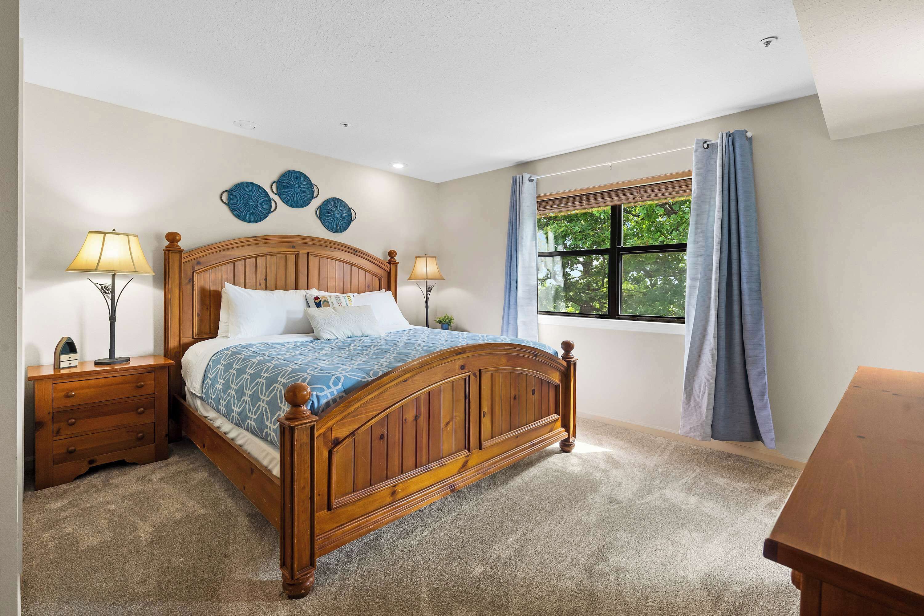 A bedroom with a wooden bed, two bedside tables, a nightstand, a window with curtains, and a view of greenery outside.