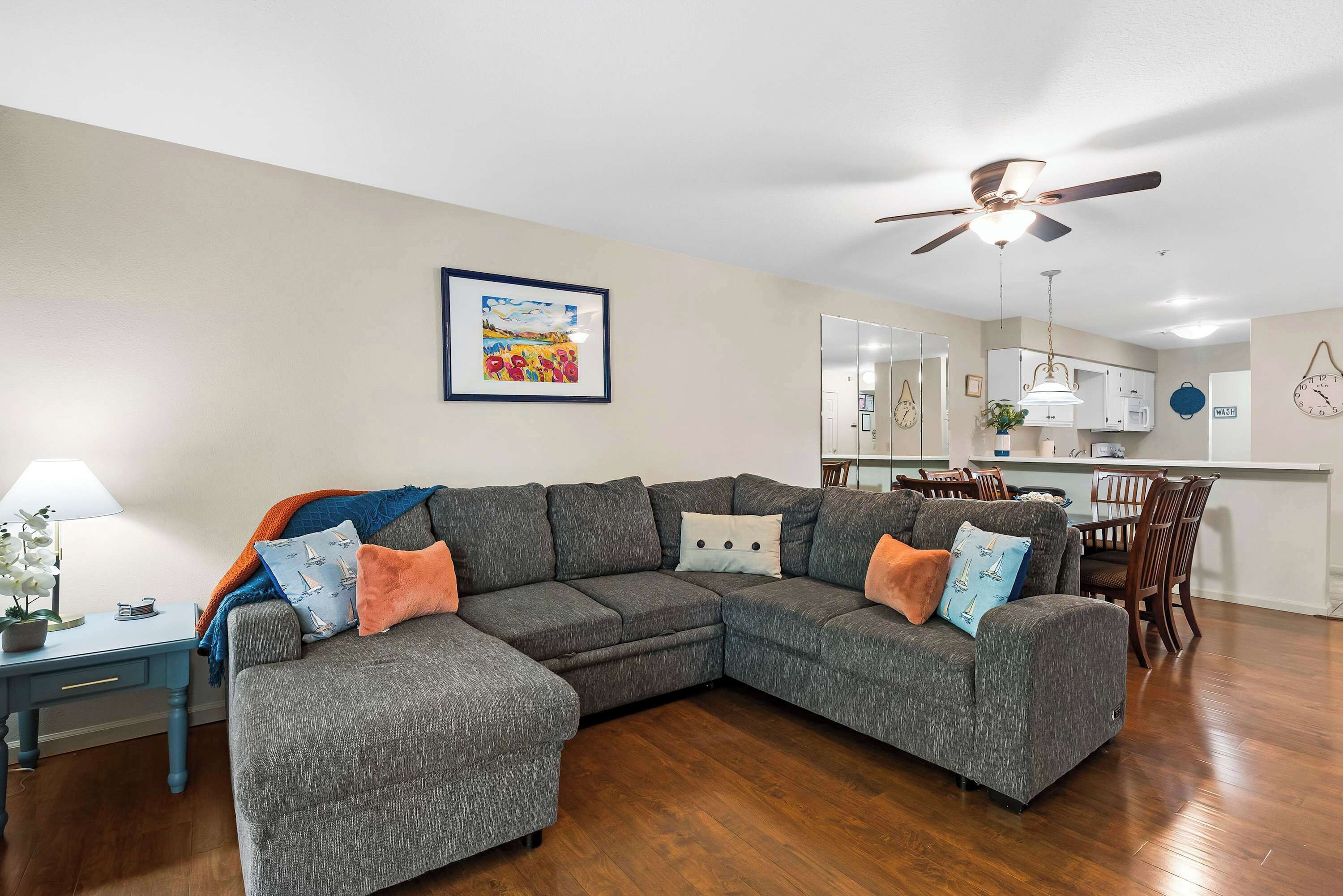 A living room with a grey sectional sofa, a small desk, a ceiling fan, and a kitchen area in the background.