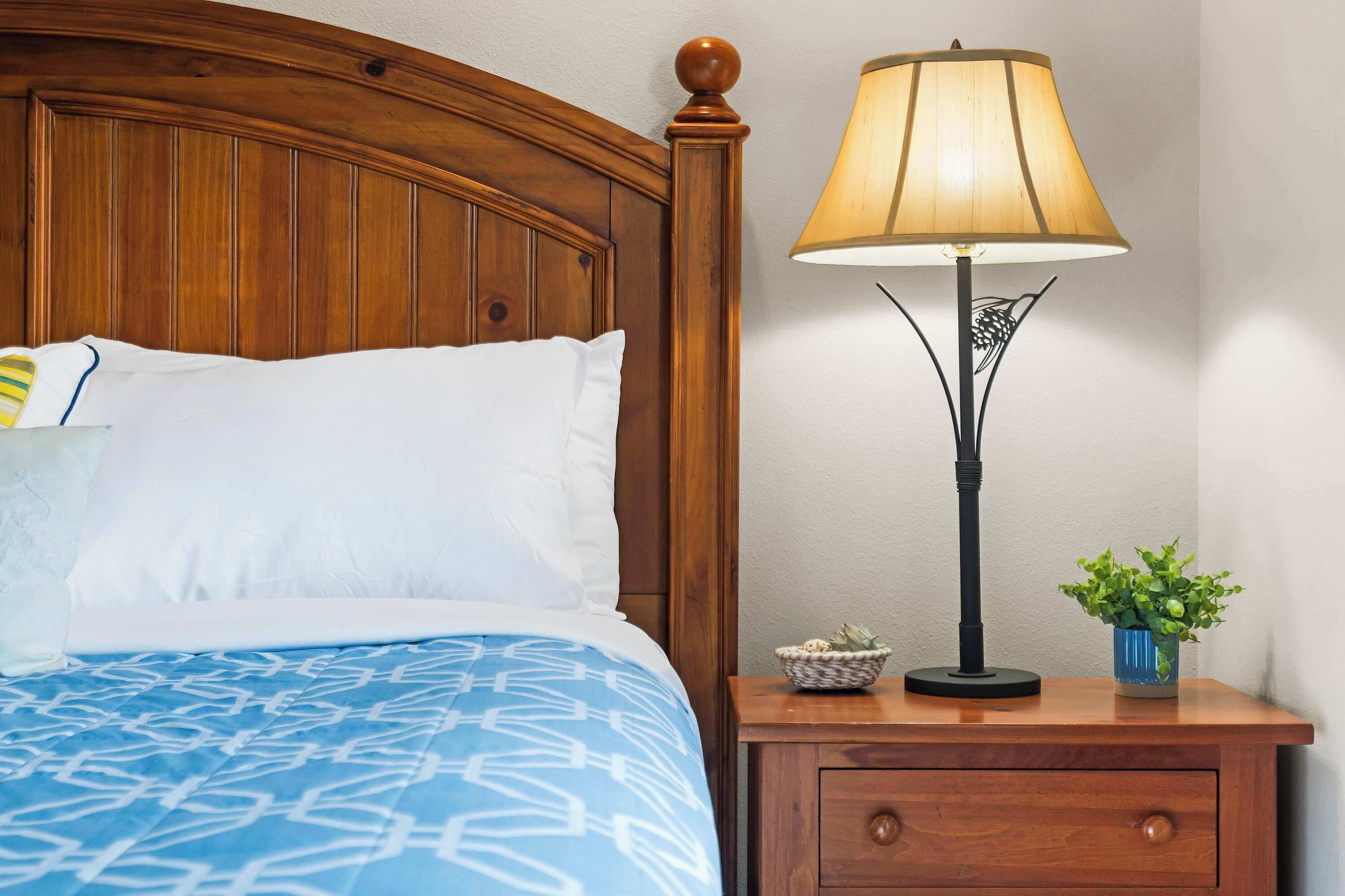 Ein ordentlich bezogenes Bett mit blauen Mustern auf der Tagesdecke und weißen Kissen, ein Nachttisch aus Holz mit einer Lampe und einer kleinen Pflanze sowie ein Holz-Kopfschild.