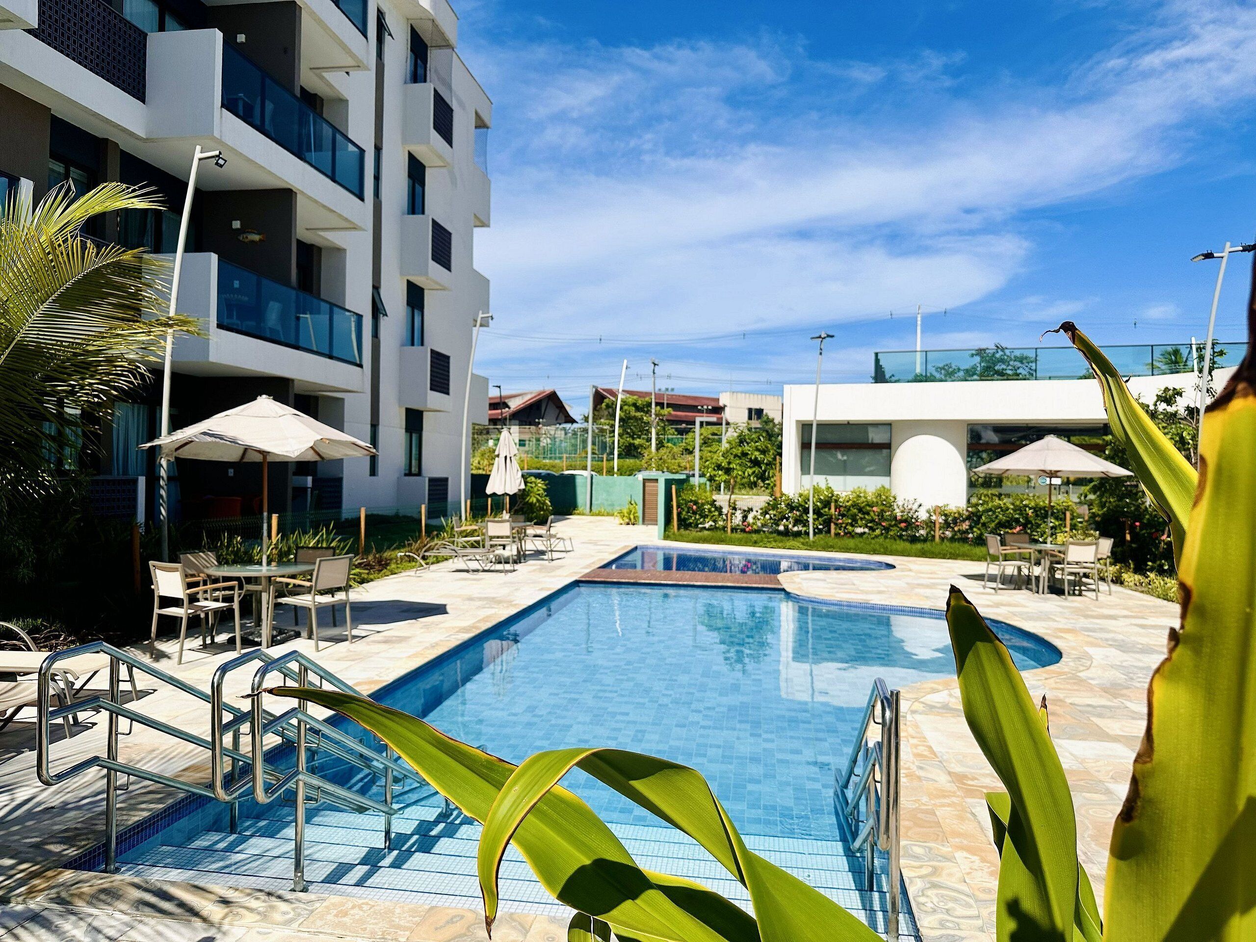 A swimming pool with steps, surrounded by outdoor seating and umbrellas, with modern buildings in the background.