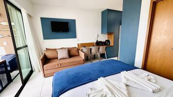A hotel room with a bed, a sofa, a desk with a chair, a television mounted on the wall, and a sliding glass door leading to a balcony.