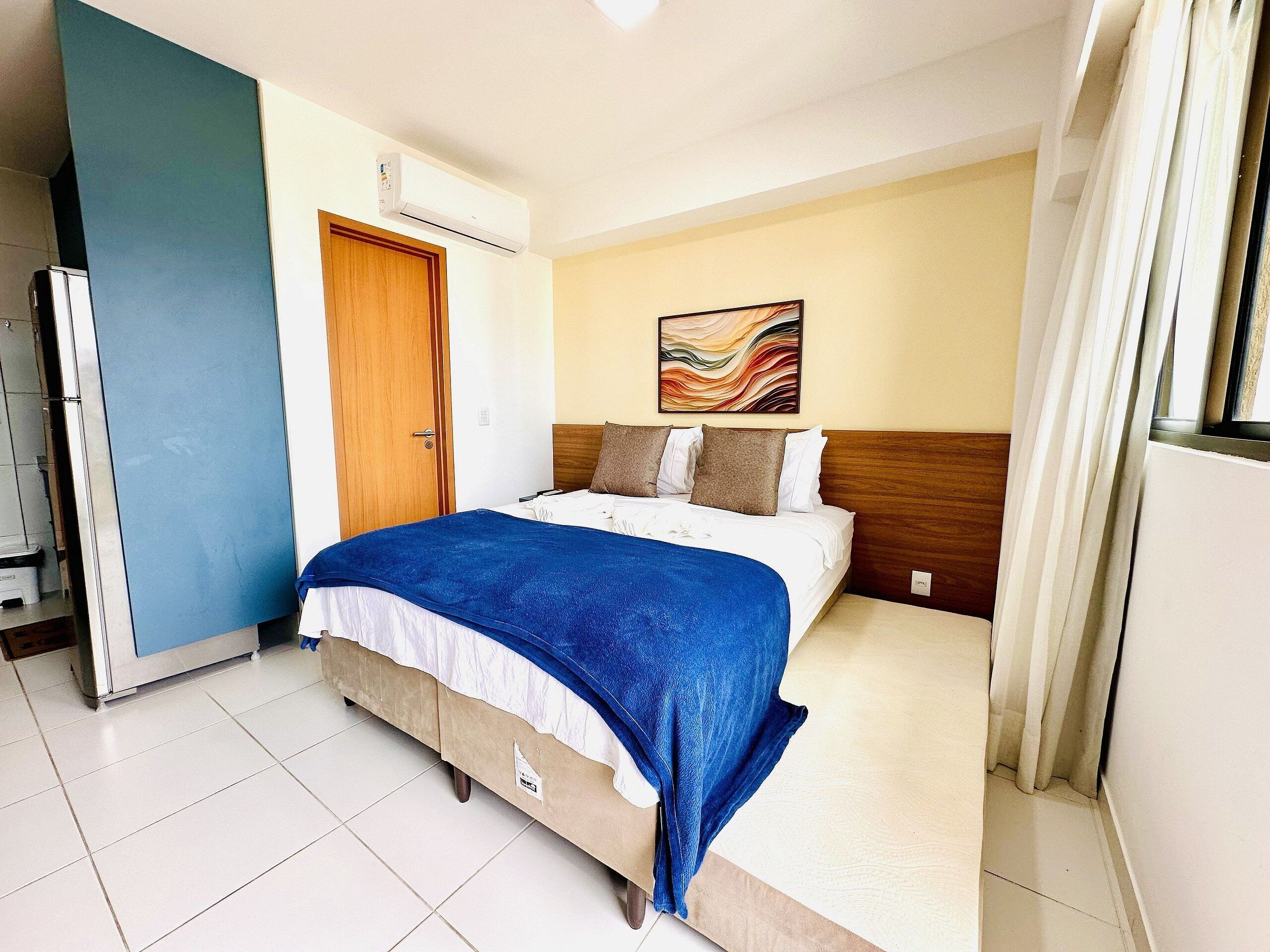 A hotel room with a bed, a door, an air conditioning unit, a painting on the wall, and a window with curtains.