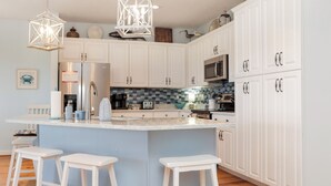 Una cocina moderna con armarios blancos, una isla central con taburetes, electrodomésticos de acero inoxidable y objetos decorativos en los estantes.