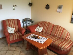 Un salon avec un canapé à rayures, un fauteuil rouge, une petite table avec un set de table, un vase et un luminaire fixé au mur.