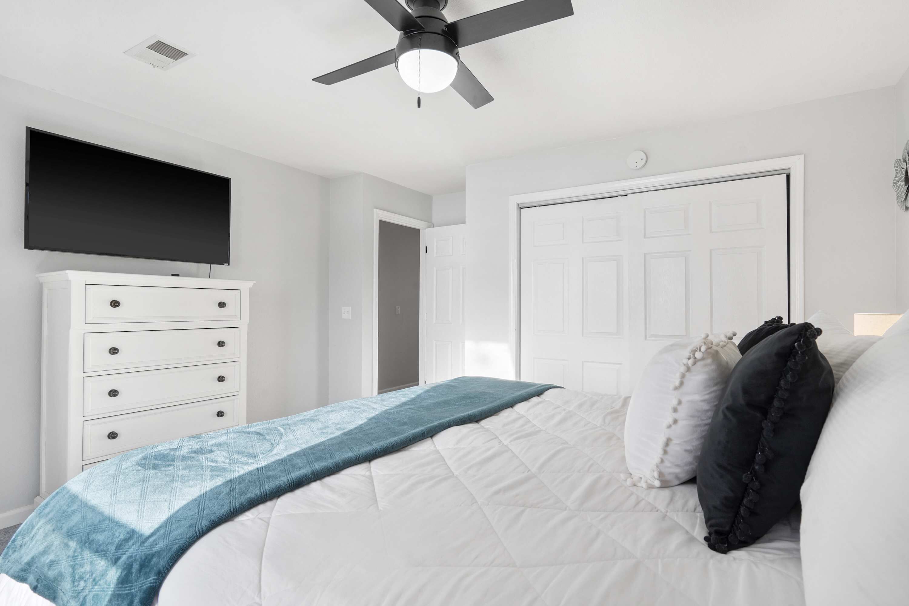 A bedroom with a bed, a dresser, a ceiling fan, a TV, and a door.