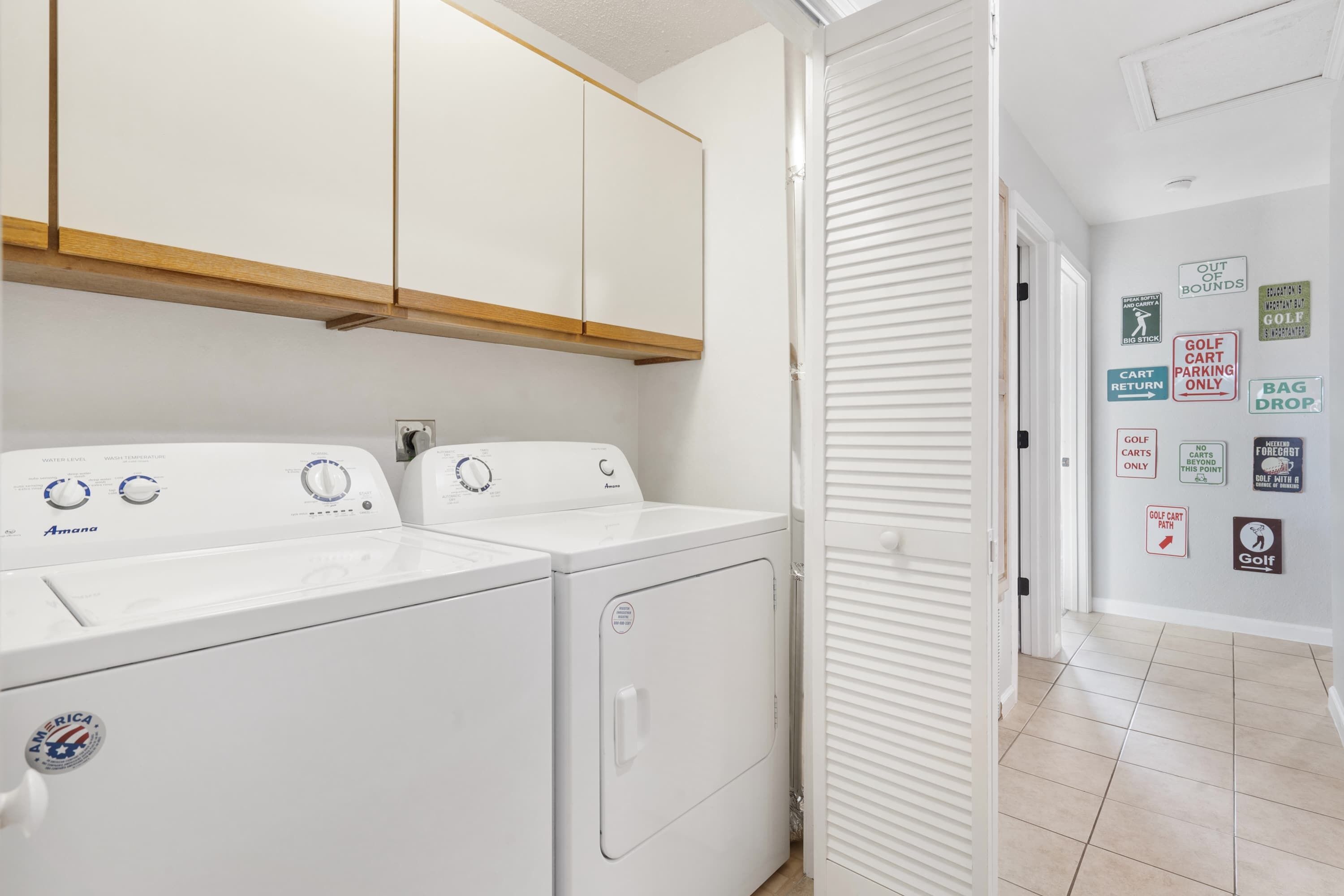 A laundry area with two white Amana washers and dryers, wooden cabinets above, and a hallway with various signs including 'GOLF CART RETURN ONLY'.