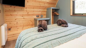Une pièce chaleureuse aux murs en bois, avec un lit recouvert d’une couverture tricotée, un téléviseur à écran plat, une petite étagère en bois et une fenêtre donnant sur un beau paysage.