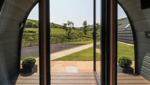 Une vue à travers une porte vitrée donnant sur une terrasse en bois avec un chemin pavé, le tout entouré d’un paysage rural avec des collines et un arbre.