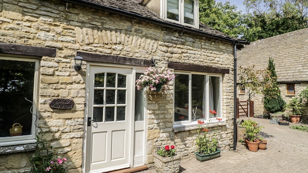 Une petite maison en pierre avec une porte blanche, une petite fenêtre ornée de fleurs et un chemin en briques.