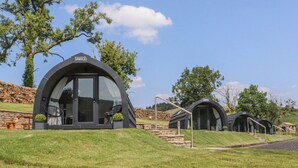 Une rangée de cabanes modernes en forme de dôme, dotées de grandes fenêtres, est installée sur une colline herbeuse, avec des murs en pierre et des arbres en arrière-plan.