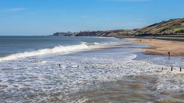 Una playa costera con olas, cielo despejado y edificios a lo lejos.