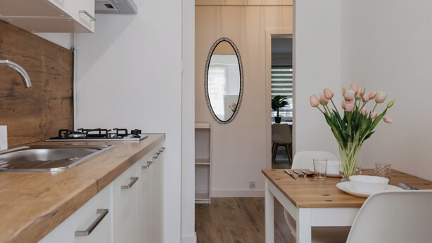 A modern kitchen with a wooden countertop, a gas stove, and a stainless steel sink. There is a dining area with a wooden table, white chairs, and a vase of flowers. A large mirror hangs on the wall above the dining table. - Apartamenty Sun & Snow Kopernika 16 (Ustka)