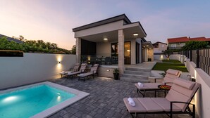 Un área de piscina moderna al aire libre con sillones de descanso y una zona de estar cubierta.
