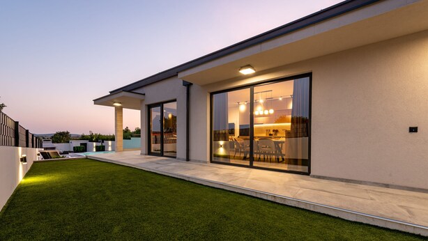 Una casa moderna con un césped bien cuidado, puertas correderas de vidrio grandes que dan a un comedor, y una cocina a la vista.