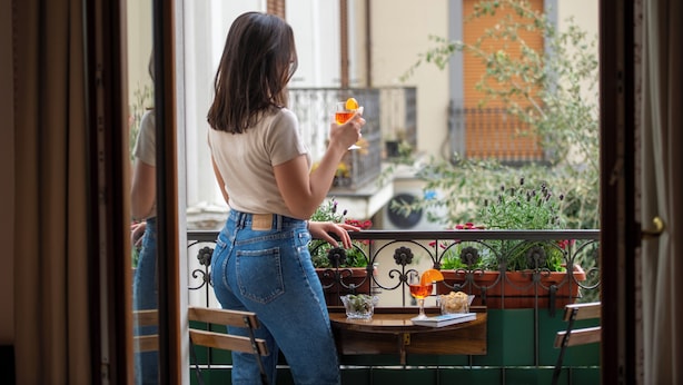 Eine Frau steht auf einem Balkon, in der Hand ein Getränk, und blickt auf einen Garten mit Topfpflanzen. Im Hintergrund ist eine Tür zu sehen.