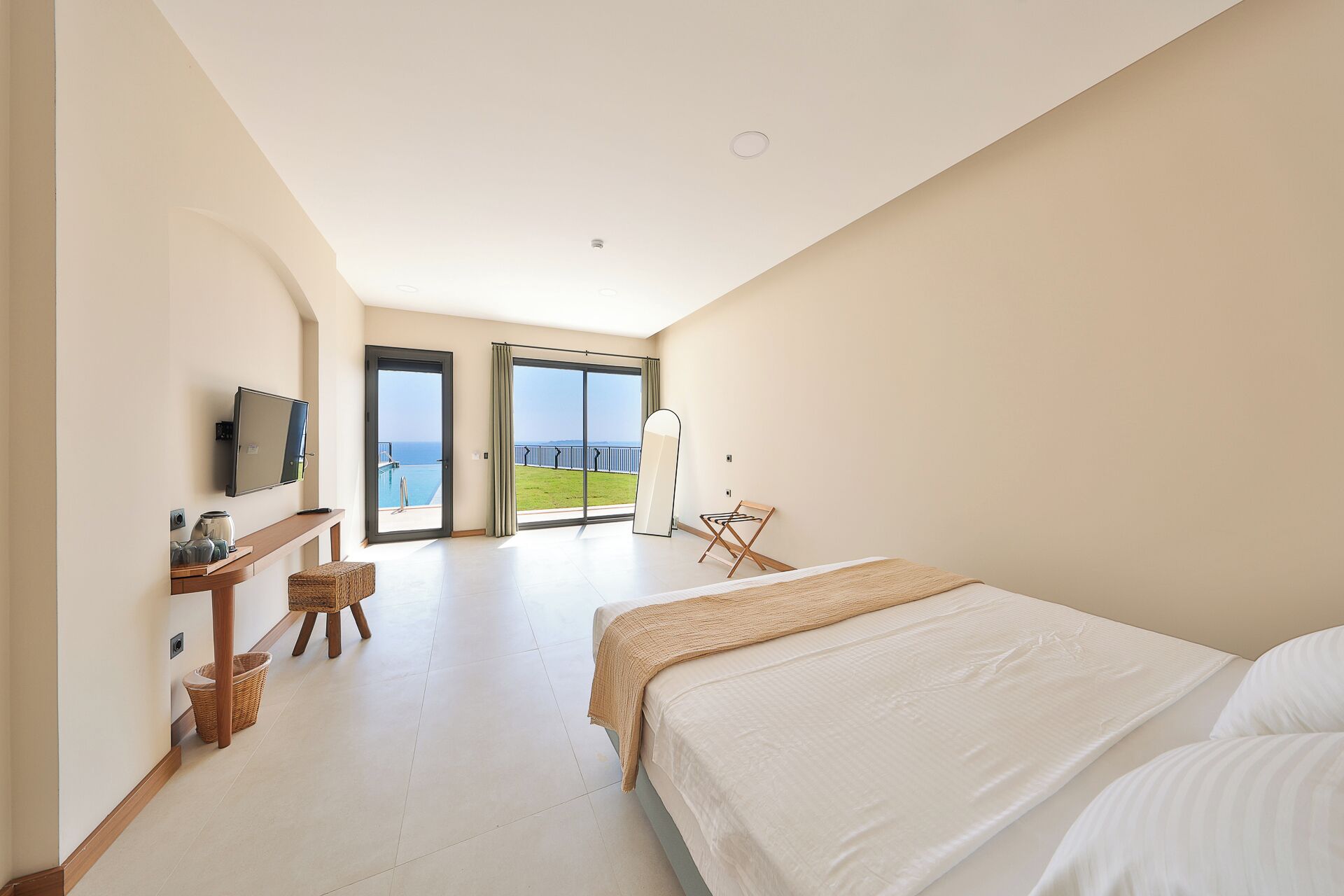 A modern hotel room with a large bed, a television, a small table with decorative items, and a view of the sea through a sliding door.