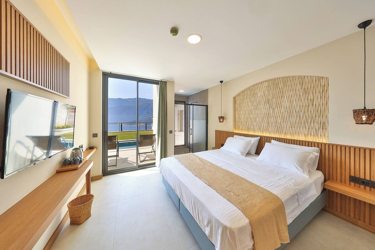 A modern bedroom with a large bed, a TV, and a balcony with a view of the sea.