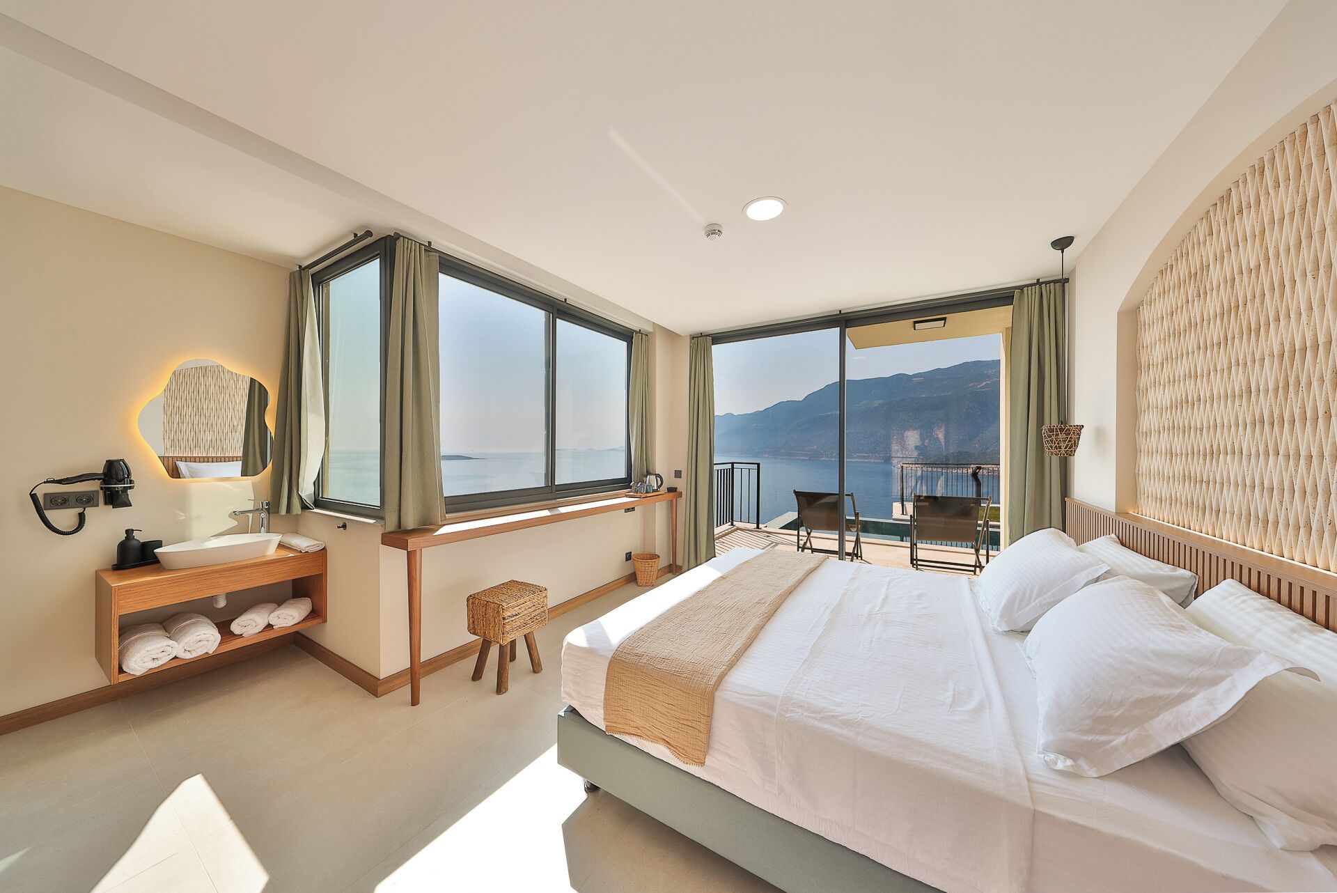 A hotel room with a bed, a desk, a mirror, a window with a view of the sea, and a balcony with a chair.