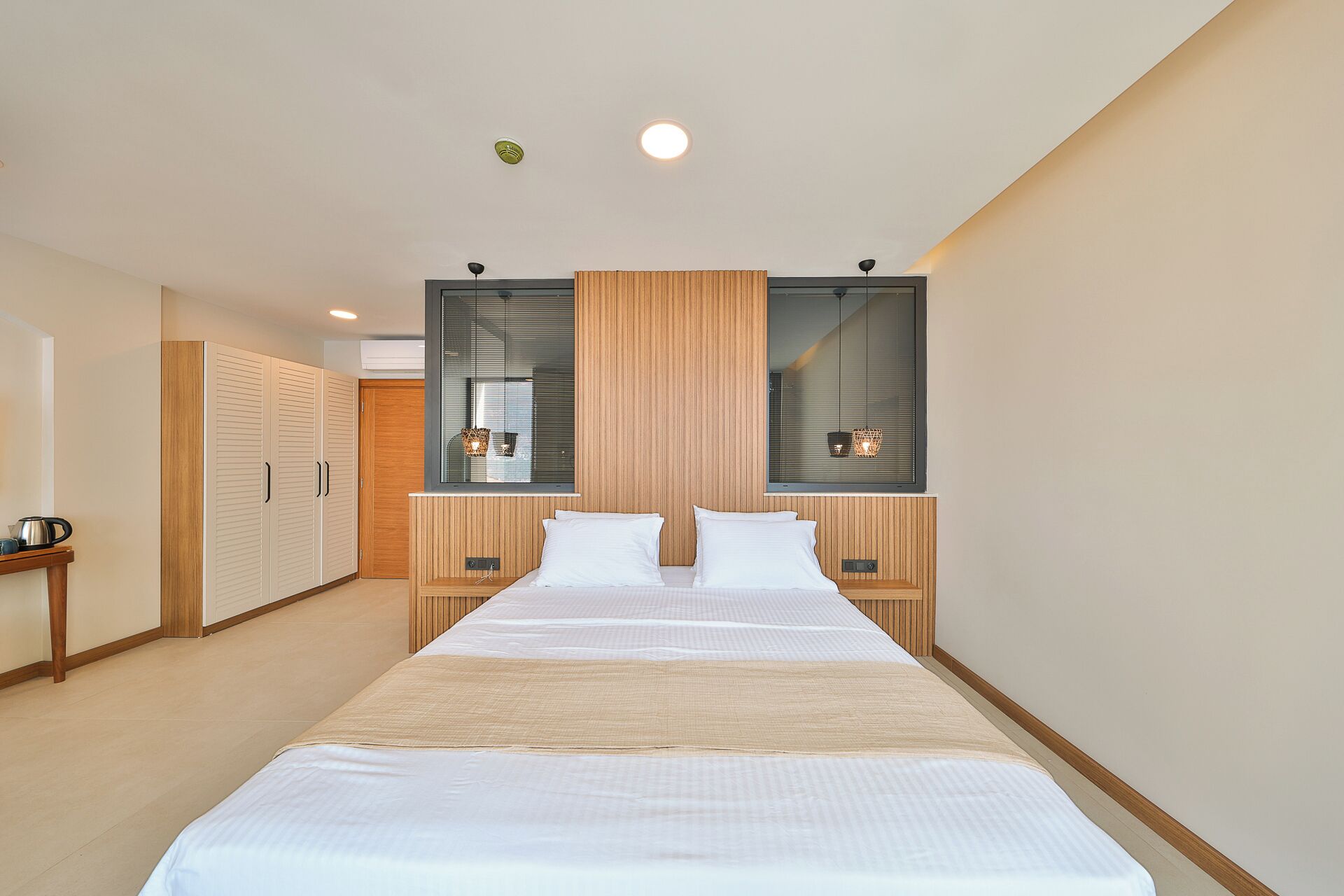 A modern bedroom with a large bed, wooden paneling, a window with blinds, and a small bedside table with a lamp.