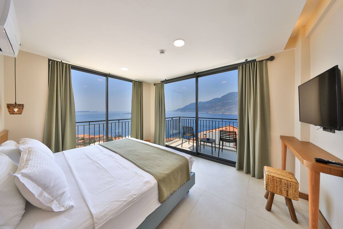 A hotel room with a large bed, a TV, a wooden desk, and a balcony with a view of the sea and mountains.