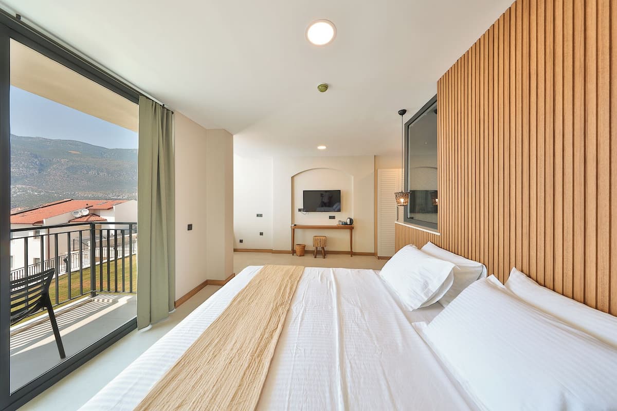 A hotel room with a large bed, a TV mounted on the wall, a small desk, and a balcony with a view of the mountains.