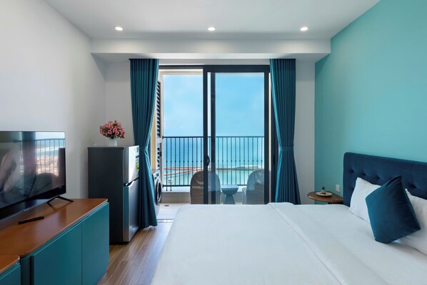 A hotel room with a bed, a TV, a refrigerator, a small table with chairs, and a balcony with a view of the sea.