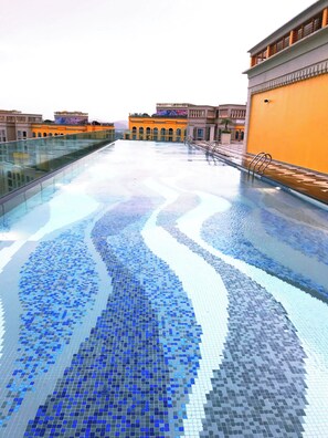 A rooftop swimming pool with a blue mosaic design, surrounded by buildings with yellow and orange facades.