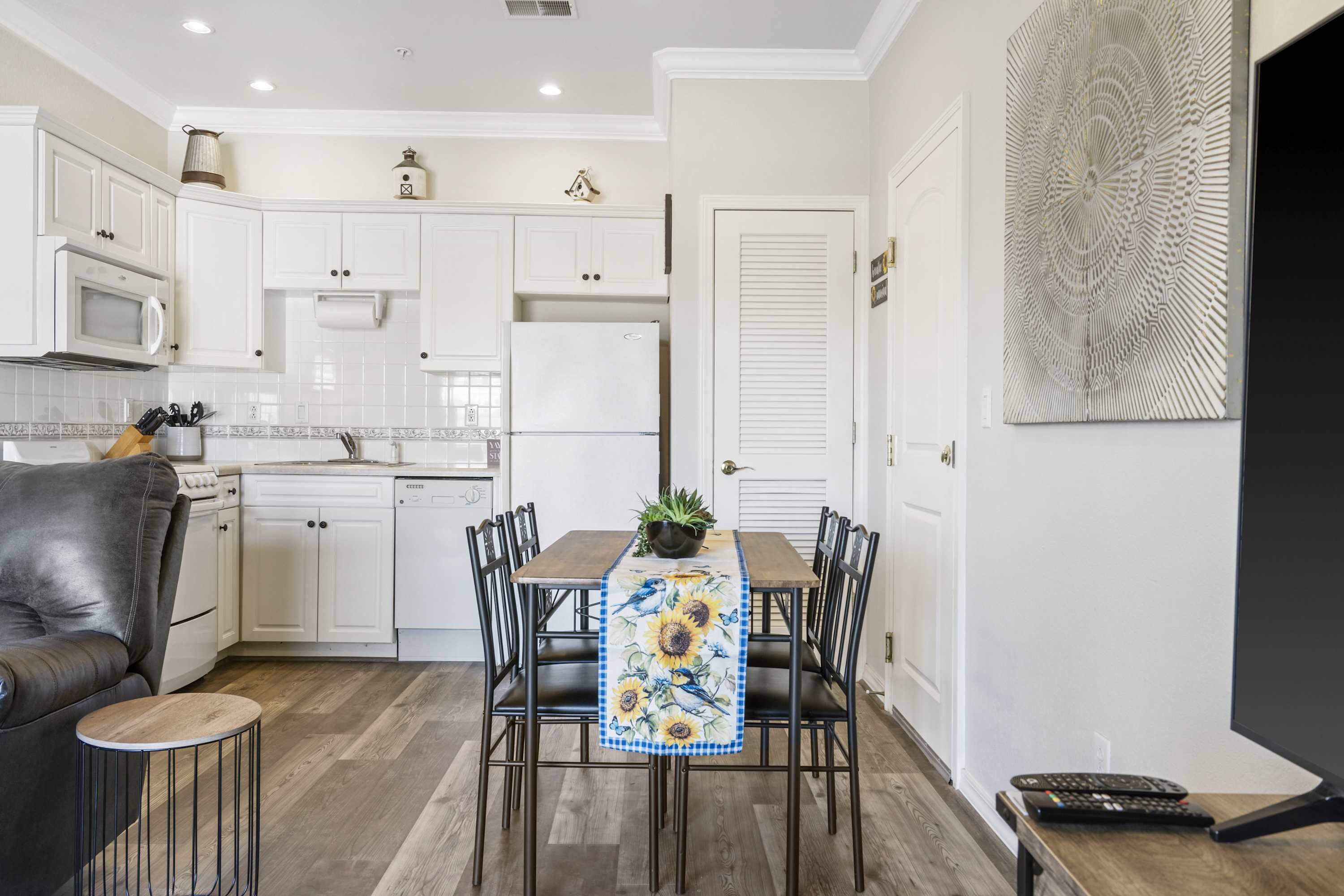 Una cocina moderna con armarios blancos, una mesa de comedor con camino de mesa de girasol y un sofá de cuero negro.
