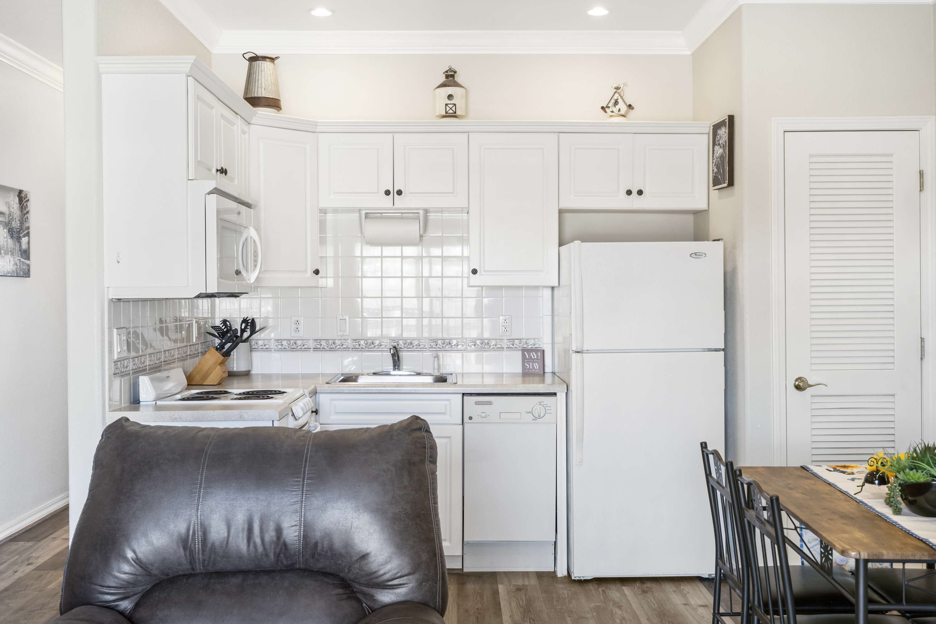 Una cocina moderna con armarios blancos, horno integrado y refrigerador. Hay una mesa de comedor con sillas y una pequeña planta en la mesa.
