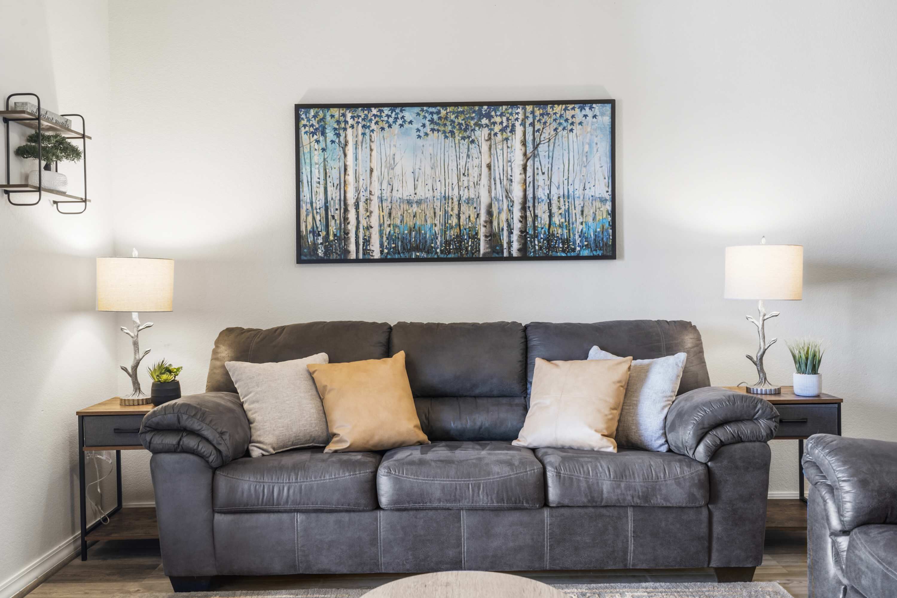 En stue med en mørk lædersofa, to sofaborde med lamper, et billede på væggen og en lille plante på et bord.